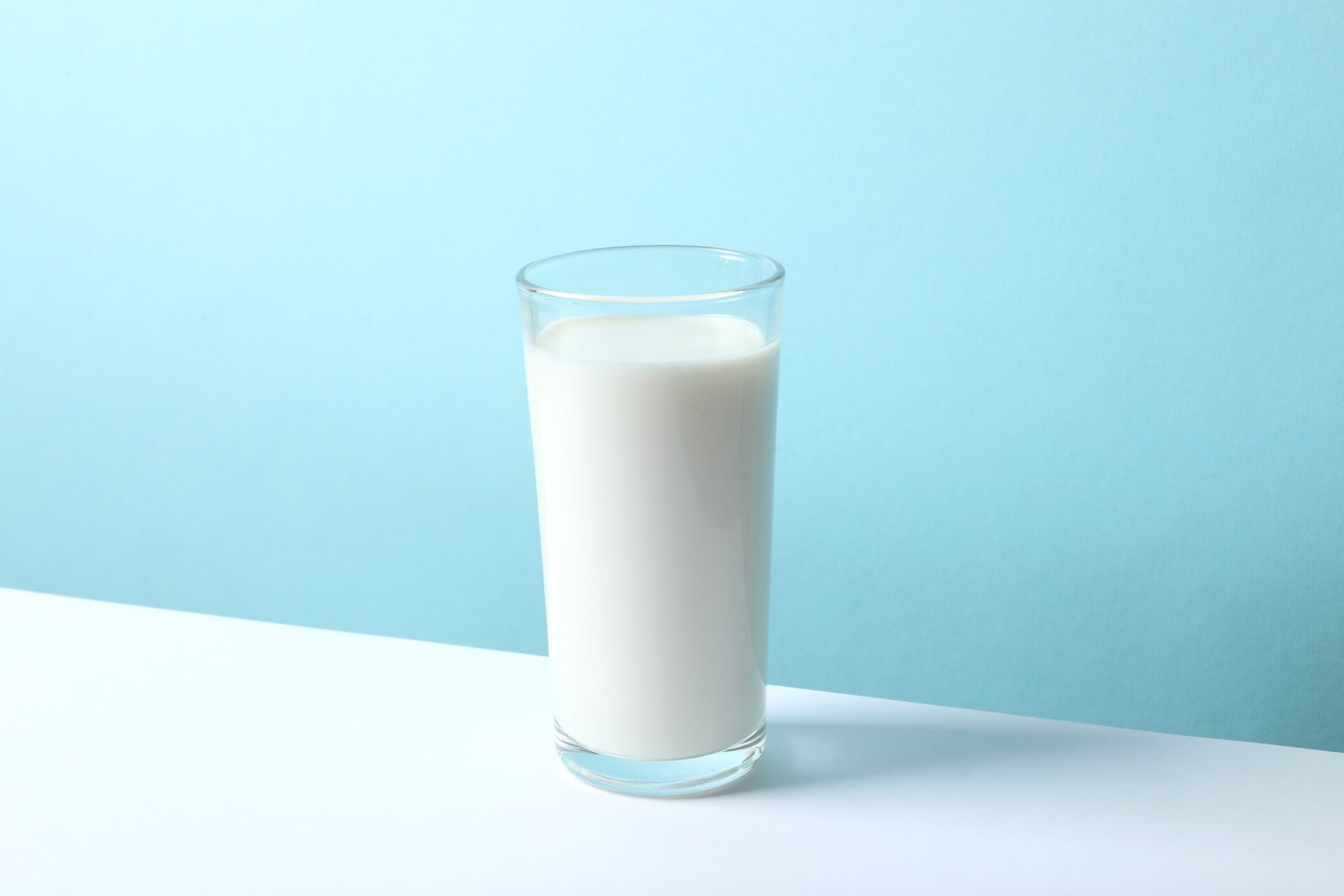 Clear glass filled with fresh white milk on a light blue background.