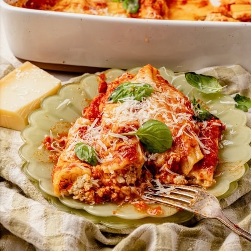 Cottage Cheese Manicotti on a plate with basil leaves on the top, a fork on the plate and in the background shows a small block of parmesan cheese